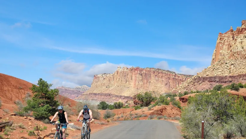 Two cyclists on road with red rocks of Capitol Reef National Park in background.