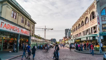 View of stone-paved street with pedestrians and "Farmers Market" neon sign on left and old buildings on right in Seattle, Washington