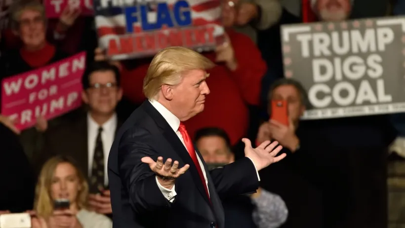 Trump at rally in Hershey, PA with person holding "Trump Digs Coal" sign visible in background.