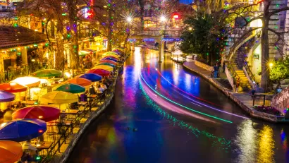 View of San Antonio River Walk with colorful umbrellas at tables on waterfront and with boat lights blurred by long-exposure