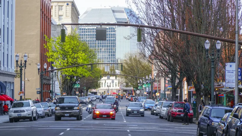A road in Portland with cars facing the camera and tall buildings on either side. The cars are stopped at a traffic light.