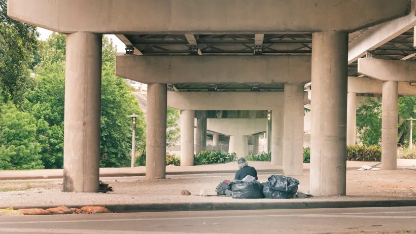 Man with grey hair sits next to black garbage bags in freeway underpass in Houston.