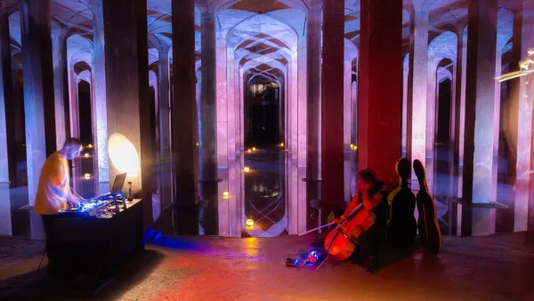 Woman cellist sitting on chair and man standing at table with music mixing equipment inside darkened cistern with concrete columns and purplish lighting.