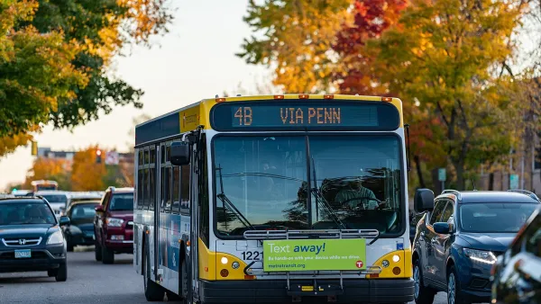 Front of city bus driving down tree-lined street, surrounded by cars.
