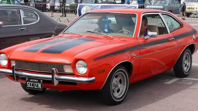 Red 1972 Ford Pinto with black racing stripes on display with man sitting in driver's seat.