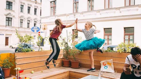 Parklet in Vienna with two young girls jumping up to high-five each other, one wearing a bright teal tutu.
