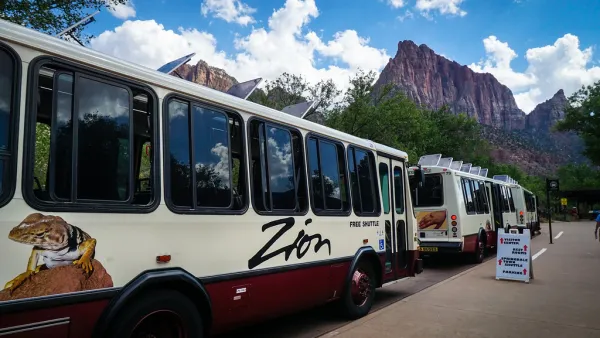 White park shuttles with large Zion logo on side and red rock cliffs in background in Zion National Park.