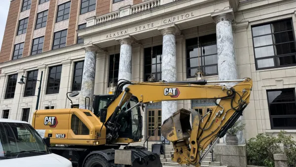 A CAT excavator drives past a brick building with marble columns.