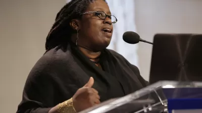 Black woman speaking at podium with presentation screen in background.