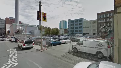 A surface parking lot, full of cars, is located between tall buildings and urban density.