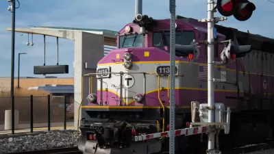 The front of a purple and gray train running along track with a rail crossing signal on the right.