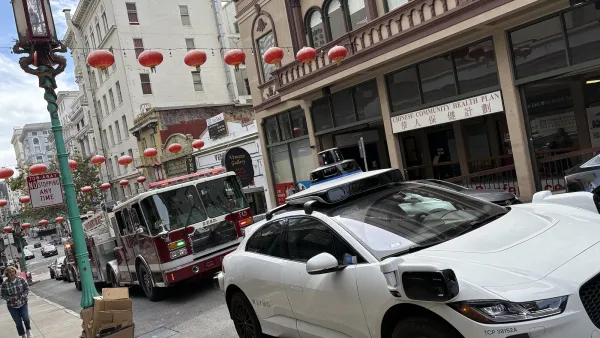 White Waymo car blocking street in front of red fire truck on narrow street with red Chinese lanterns in Chinatown, San Francisco, CA.
