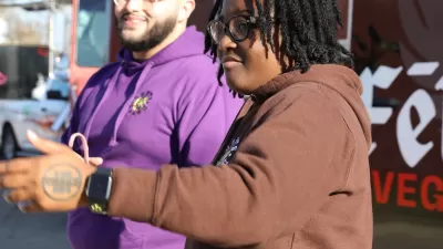 Black woman in brown sweatershirt and Black man in purple sweatshirt stand in front of food truck.