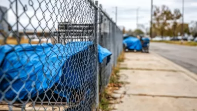 Blue tarps covering tents set up by unhoused people along chain link fence on concrete sidewalk.