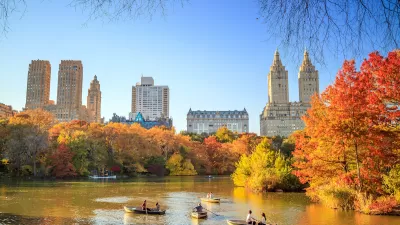View of lake in Central Park, New York City with people in rowboats and bright red and orange fall trees, buildings in background.