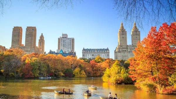 View of lake in Central Park, New York City with people in rowboats and bright red and orange fall trees, buildings in background.