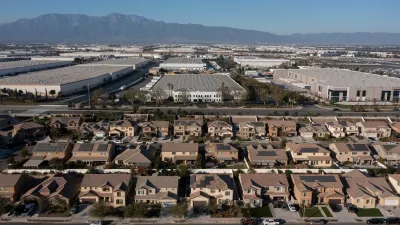 Aerial view of large warehouses across from development of suburban single-family homes in Jurupa, California with desert mountains in background.