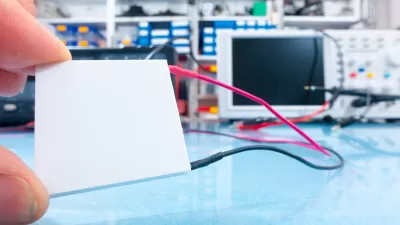 Close-up of hand holding small white square thermoelectric generator in lab with computer in background.