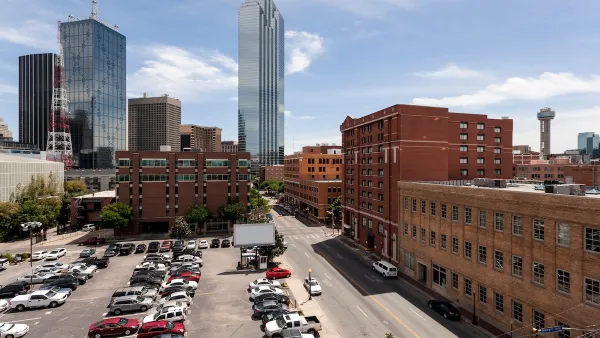 View of mostly full parking lot next to multi-story red brick buildings with modern glass skyscraper in background in downtown Dallas, Texas.