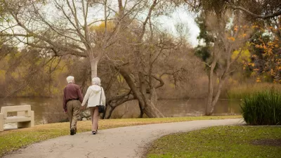 View from back of elderly man and woman holding hands and walking on a path in a park with fall colors.
