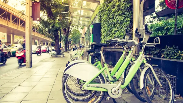 Green bike share bikes parked in a row on a commercial street with outdoor dining and greenery.