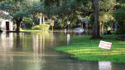 Flooded residential street with houses, yards, and trees on each side, a yard sign that reads "high water, no outlet," and a flooded car in the distance.