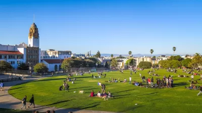 View of people sitting in lightly sloped green lawn with Spanish colonial-style building and cityscape in background in San Francisco, California.