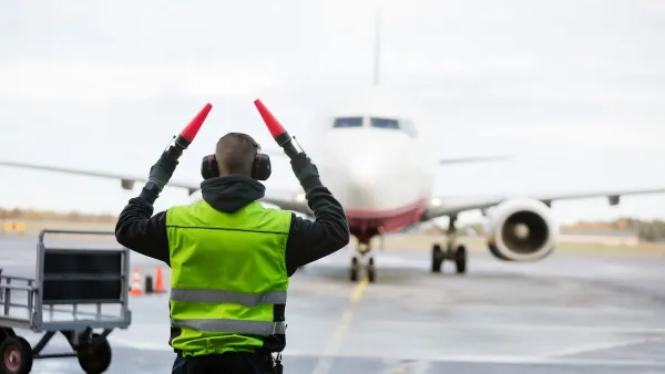 Ground crew person signaling to airplane with red batons on runway on a rainy day.
