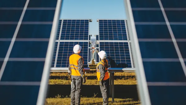 Two workers in white hard hats looking up at large photovoltaic solar panels.