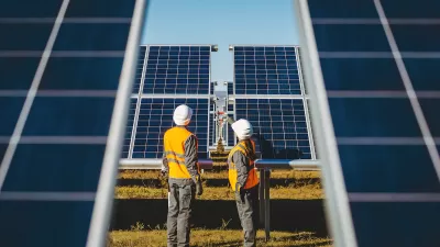 Two workers in white hard hats looking up at large photovoltaic solar panels.