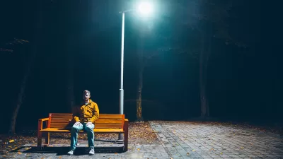 Man sitting on wooden bench under tall streetlight at night.