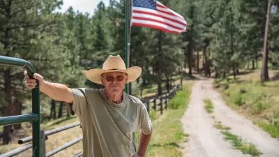A cowboy rests his hand on a green gate on a sunny day in Colorado. The United States flag is behind him and he is wearing a straw cowboy hat and glasses. A dirt road is next to him.