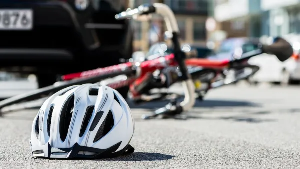 Close-up on white bike helmet lying on pavement with blurred red bike on its side in background abd black car visible behind it.