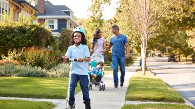 African-American parents push stroller down residential neighborhood sidewalk as young child rides scooter in front of them.