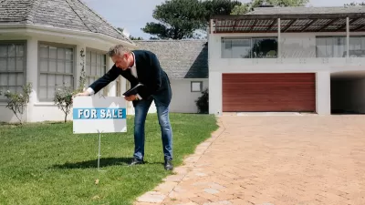 Real estate agent wearing jeans and sport coat planting For Sale sign on front lawn of home.