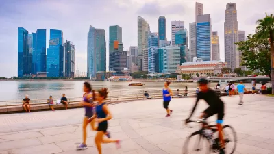 People running and cycling on wide paved path next to water with Singapore skyline with high-rises in background.