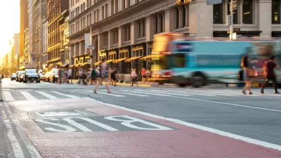 Low shot of red painted bus lane on New York City street with blurred bus, pedestrians, and buildings in background.