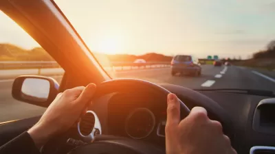 Cose-up of person's hands on steering wheel driving down highway with sunset sky.