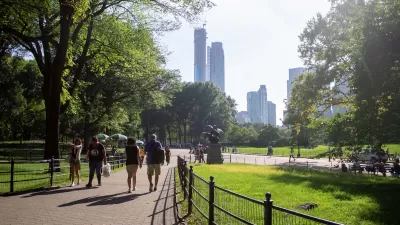 People walking on paved path in green city park with trees and tall city skyscrapers in background.