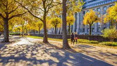 Two women walking on wide walkway among large trees next to neoclassical building in Cologne, Germany.