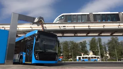 Blue electric bus at charging station with monorail train passing on elevated rail above and older model trolley bus in background.