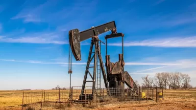 Black oil well pumpjack in brown field surrounded by metal fence.