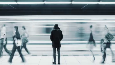 Monotone photo of man waiting on subway platform with blurry train passing by and other passengers walking by