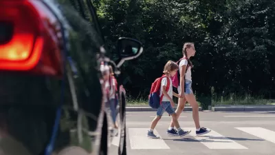 Two young children crossing street at a crosswalk in front of black car.