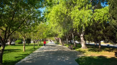 Paved walkway with a few people in lush green city park with large trees and benches.