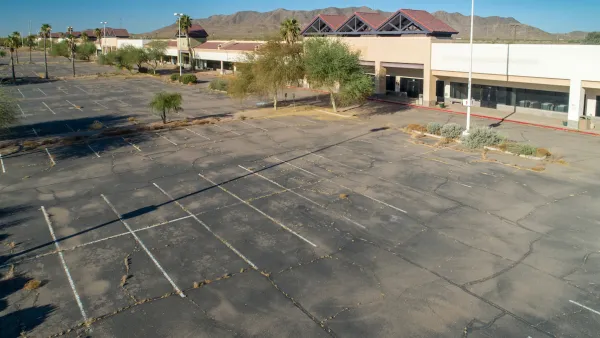 Vacant mall with vast empty parking lot with cracked asphalt and Southwestern mountains in background.