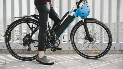 Close-up of black electric bike with person resting one foot on ground and holding blue helmet.