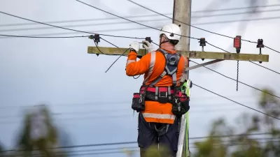 Worker in orange shirt and white helmet works at top of power line.