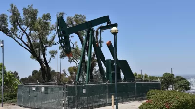 Green oil well pump jack surrounded by fence on a hill overlooking neighborhood.