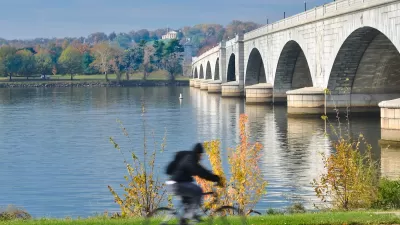 Person on bike on path next to Memorial Bridge in Washington, D.C. with fall foliage in background across river.